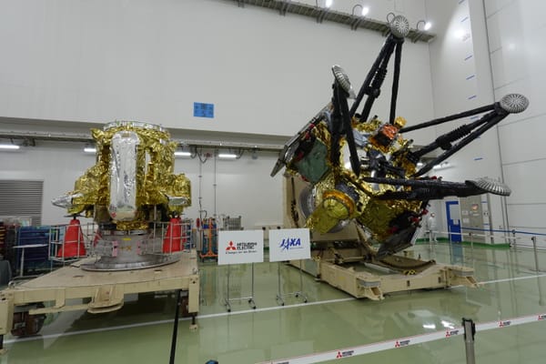 Two MMX spacecraft modules, one wrapped in gold foil and one with four landing legs, on carts in a white clean room